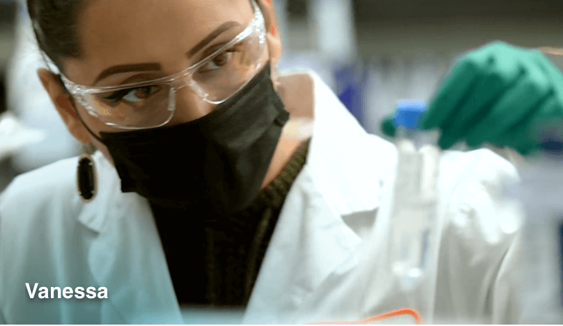 Person in protective eyewear, face mask & lab coat holding test tube
